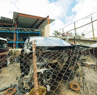 A damaged car is positioned behind a rusty chain-link fence in a scrapyard setting. Surrounding the car are various automotive parts scattered on the ground. In the background, partially visible vehicles and metal shelving filled with disassembled car components can be seen. The area is enclosed by a metal fence, and a small structure with a corrugated metal roof is nearby.